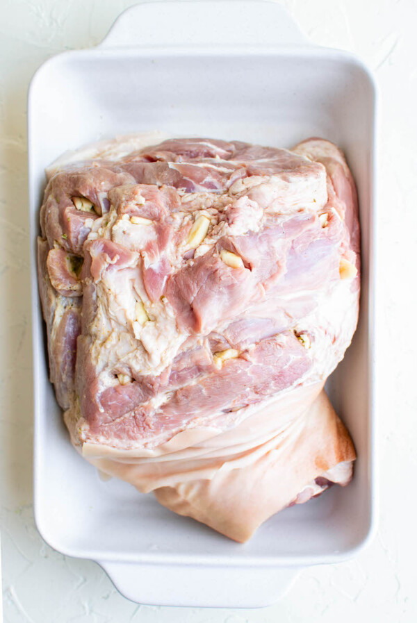 A raw cut of pork sits in a white baking dish stuffed with garlic.