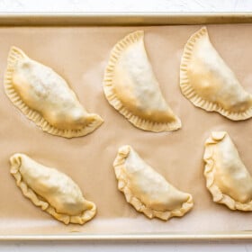 Six unbaked empanadas on parchment paper on a cookie sheet.