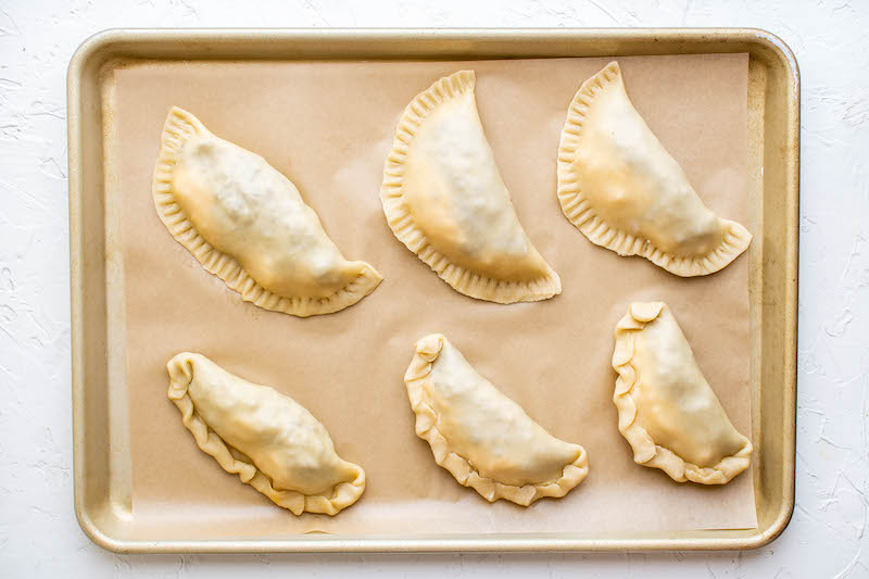 Six unbaked empanadas on parchment paper on a cookie sheet.