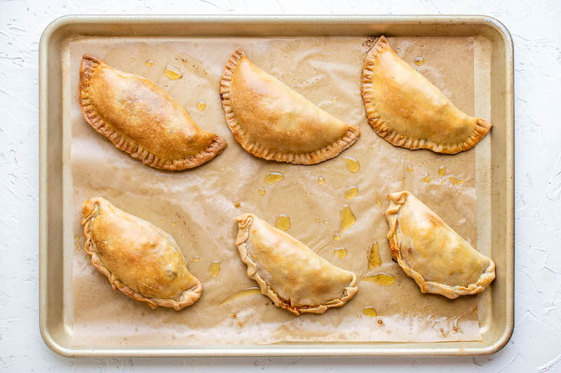 Overhead image of 6 baked empanadas on a cookie sheet with parchment paper.