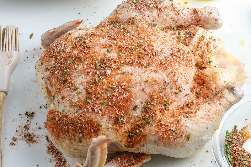 Raw whole chicken on a cutting board with seasonings on top.