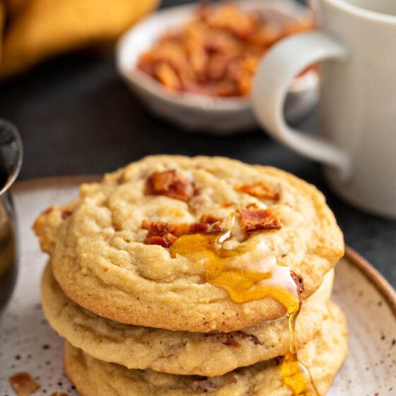 A stack of pancake cookies sits on a white plate, drizzled with maple syrup.
