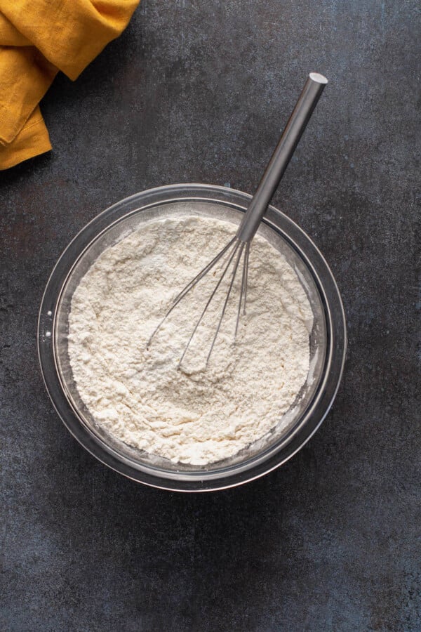 A whisk sits in a glass bowl of white dry ingredients. 