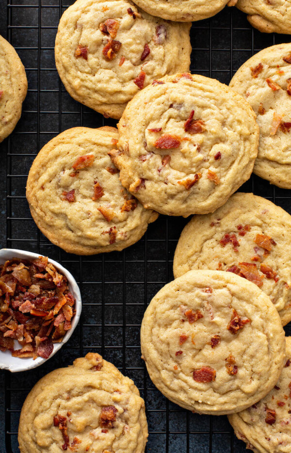 Several cookies are placed next to a small cup of bacon on a dark surface.