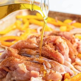 Oil being drizzled over raw chicken, peppers and onions on a baking sheet with fajita seasoning