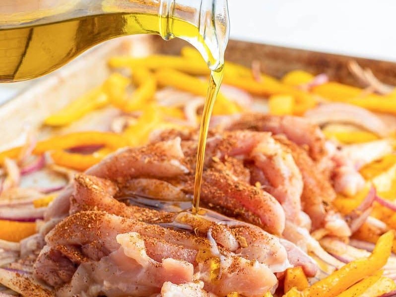 Oil being drizzled over raw chicken, peppers and onions on a baking sheet with fajita seasoning