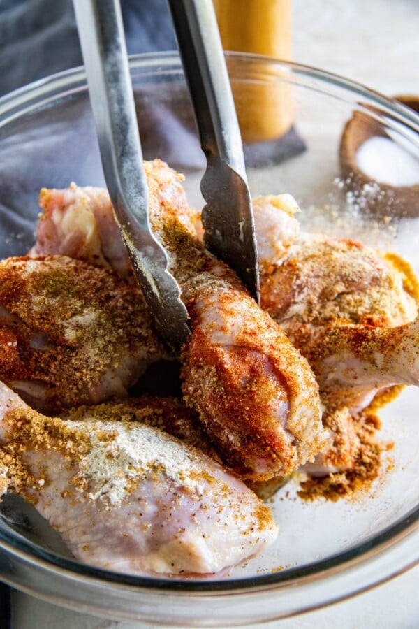 Chicken legs are being seasoned in a glass mixing bowl. 