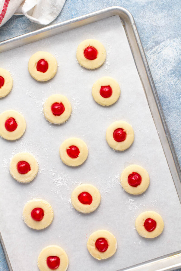 Maraschino cherries are pressed into the cookie dough balls on a cookie sheet.