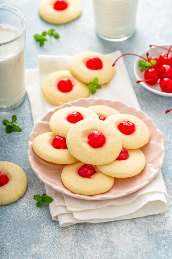 A pink plate filled with shortbread cookies with cherries on a white napkin with glasses of milk.