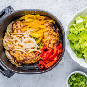 Overhead image of an air fryer basket with roasted vegetables placed on top of chicken breasts and a bowl of romaine lettuce on the side.