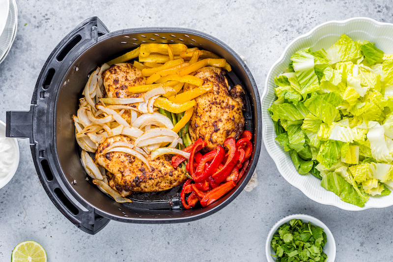 Overhead image of an air fryer basket with roasted vegetables placed on top of chicken breasts and a bowl of romaine lettuce on the side.
