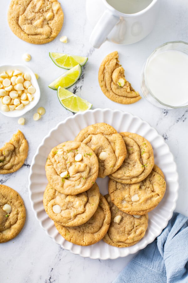 A plate filled with White Chocolate Key Lime Cookies is placed next to several other cookies on a white surface. 