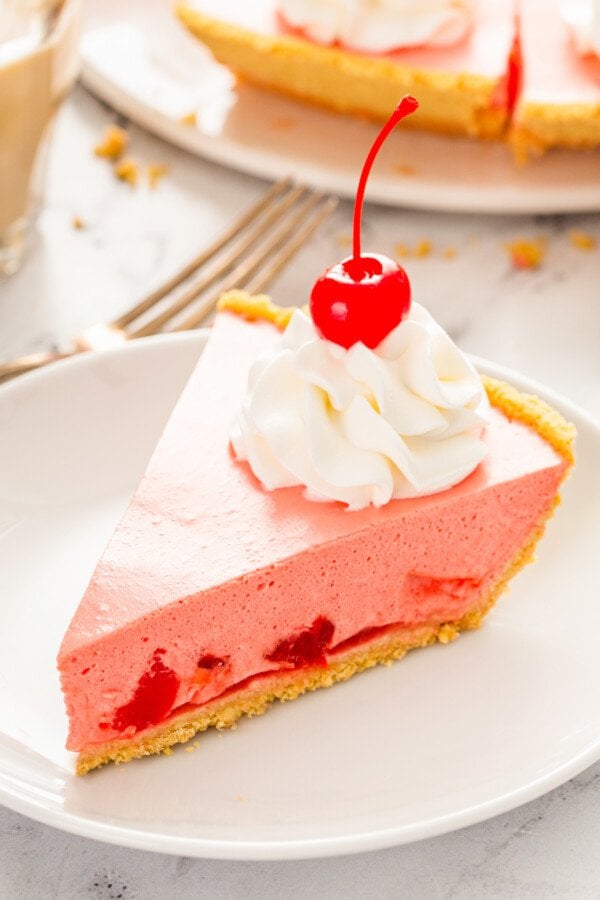 A slice of no bake cherry pie on a white plate with whip cream and a cherry on top.