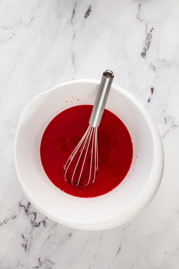 A white bowl with cherry jello in it and a whisk.
