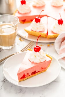 A slice of cherry jello pie on a white plate with the full pie behind it in the background.