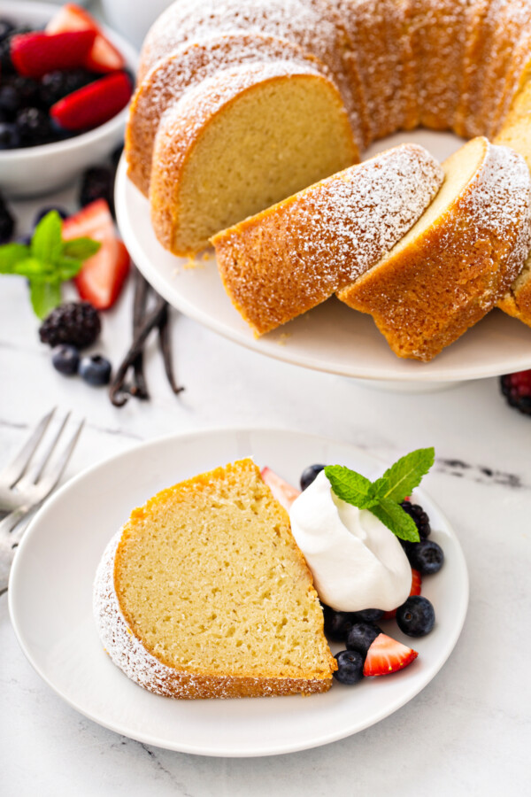 A slice of vanilla bean pound cake on a white plate with whip cream and fresh berries. 