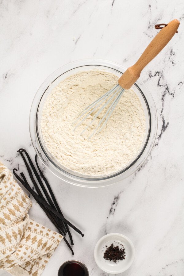 Flour in a clear glass bowl with a whisk and vanilla beans next to it.