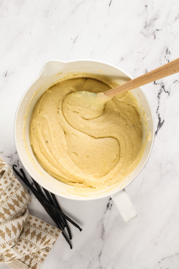 Overhead image of a mixing bowl with pound cake batter inside with a spatula and vanilla beans and a tea towel next to the bowl.