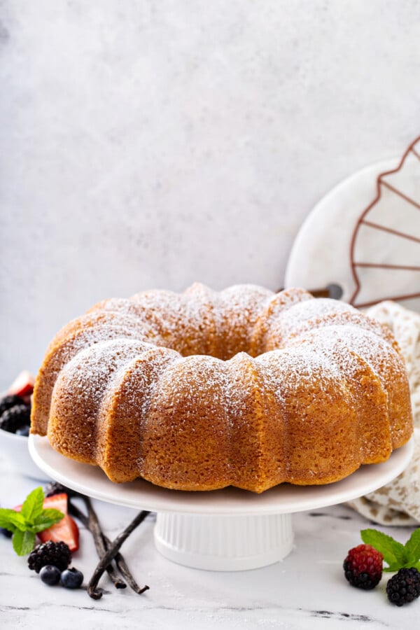 Side view of vanilla pound cake on a cake stand with powdered sugar sprinkled on top and vanilla beans next to it. 