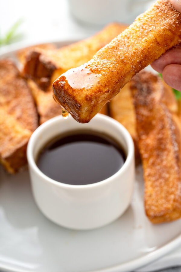 A French toast stick is being dipped into a small cup of syrup. 