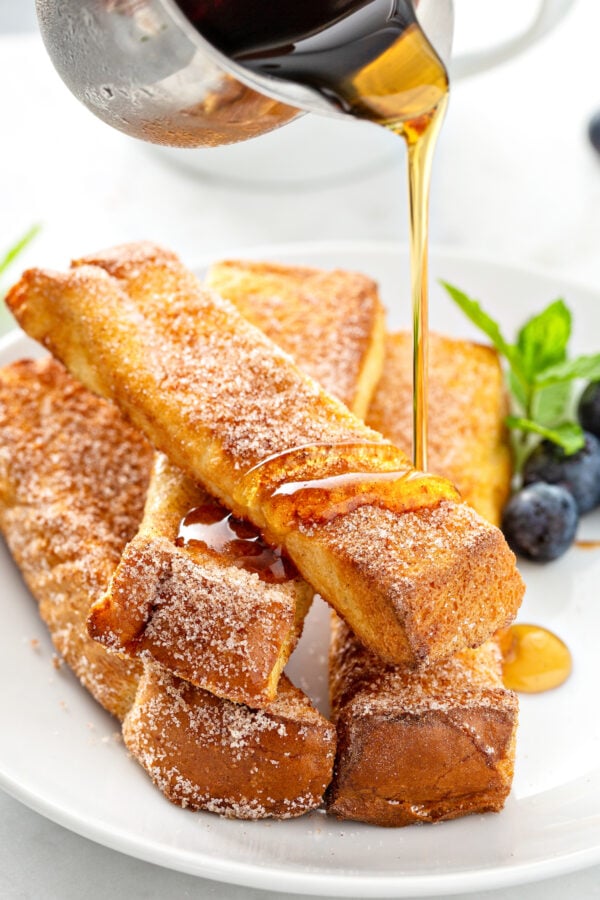 syrup is being poured over a stack of French toast sticks on a white plate. 