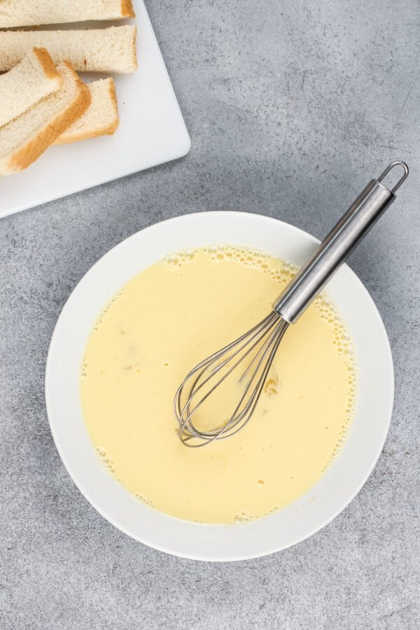 A bowl of milk, eggs, and vanilla is mixed with a whisk.