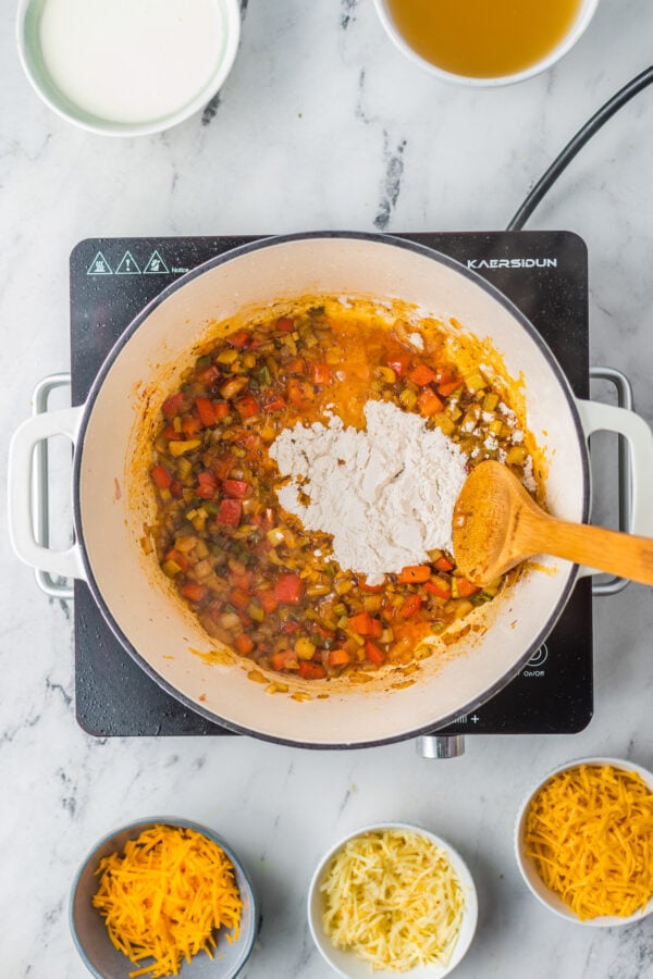 Sautéed vegetables in a large pot are being stirred with flour. 