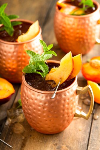 Three copper mugs are filled with peach Moscow mules.