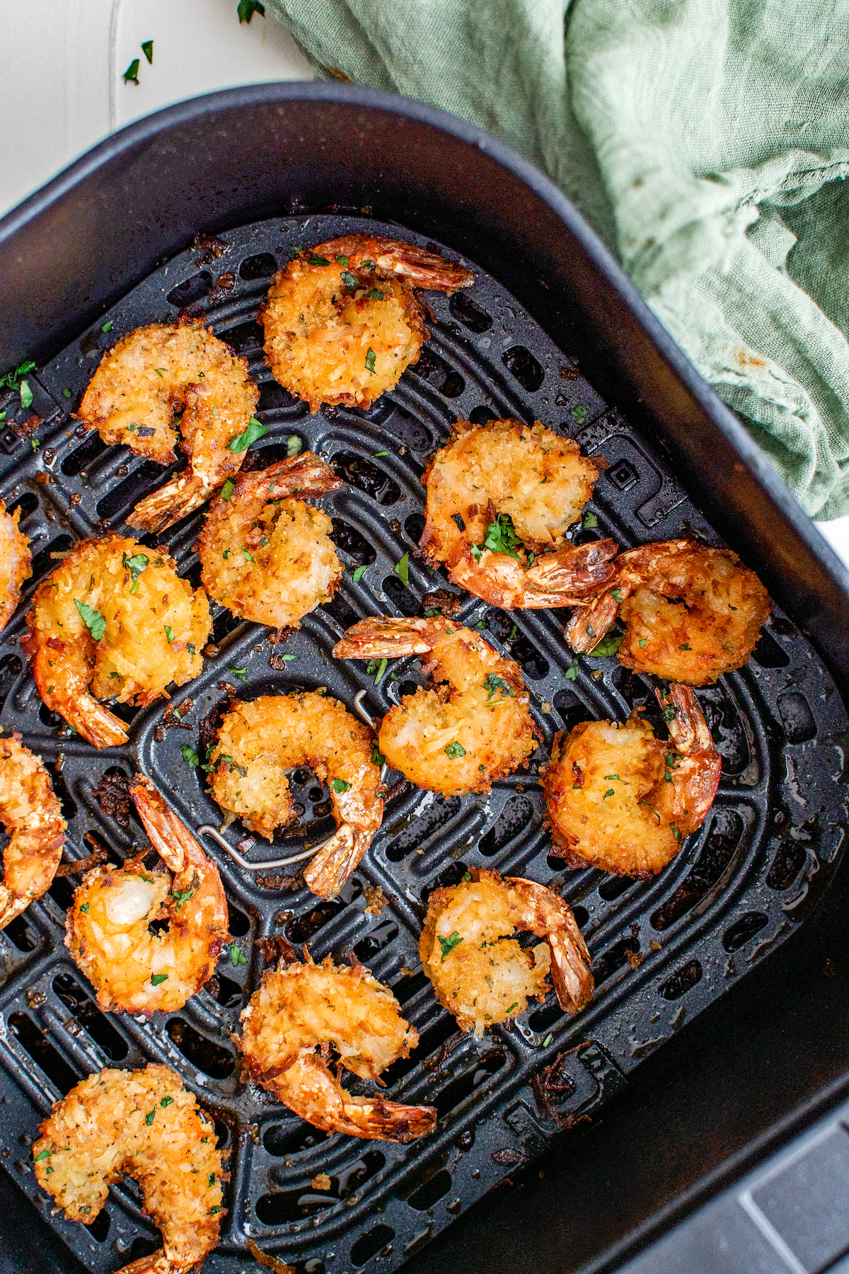 Crispy, golden brown coconut shrimp in air fryer basket after cooking topped with fresh parsley.