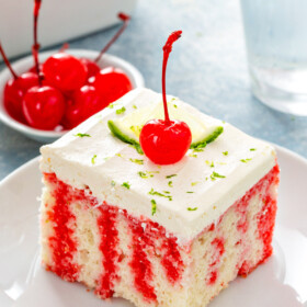 A slice of cherry limeade cake on a white plate with lime and a cherry on top.