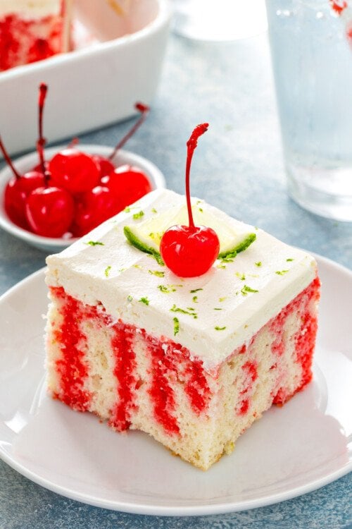 A slice of cherry limeade cake on a white plate with lime and a cherry on top.