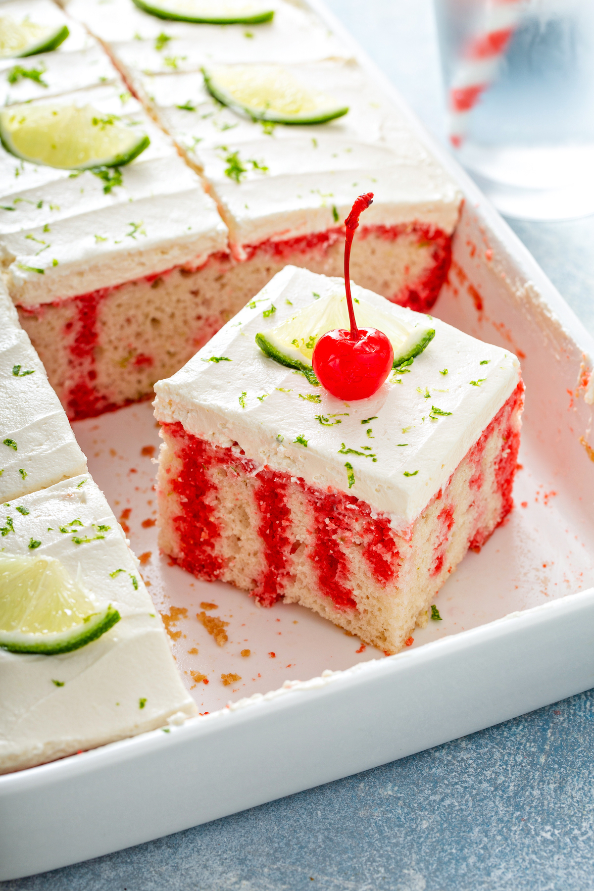 A slice of cherry limeade poke cake in the baking pan with lime zest and a cherry on top. 