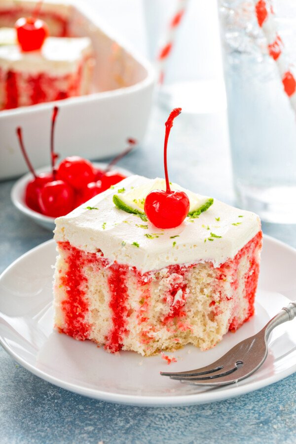 A slice of cherry limeade cake on a white plate with lime and a cherry on top and a fork taking a bite out of the cake. 