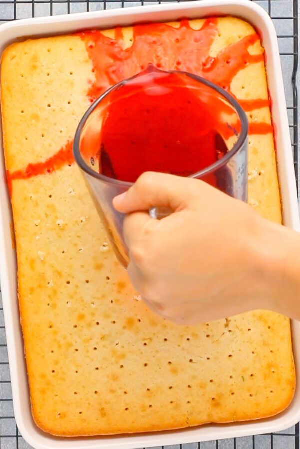 Red jello being poured over a white poke cake. 