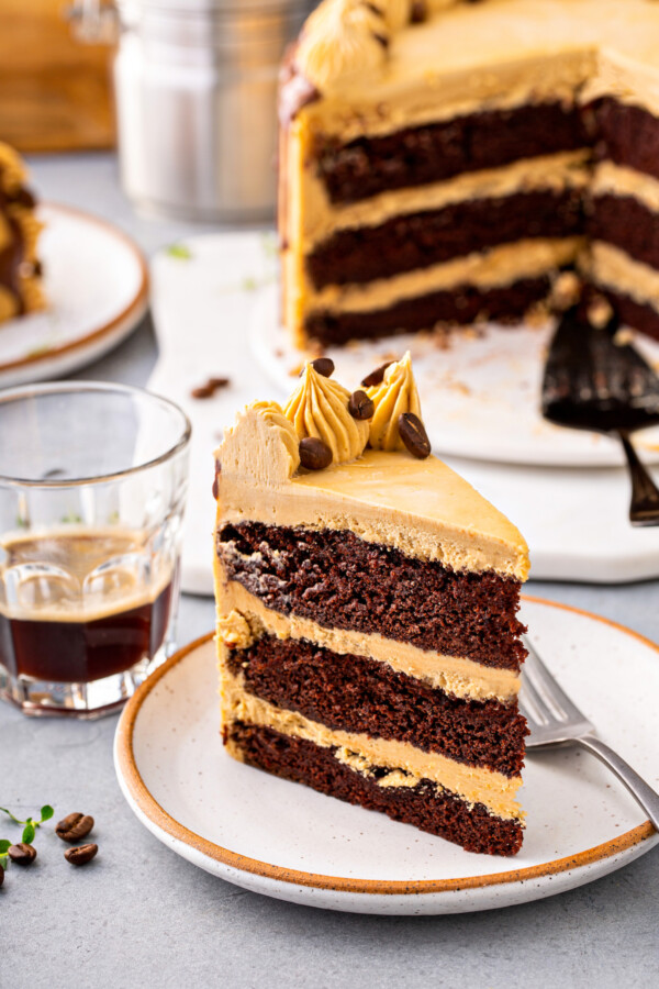 A chocolate layer cake is presented on a white plate with a small cup of espresso next to it. 