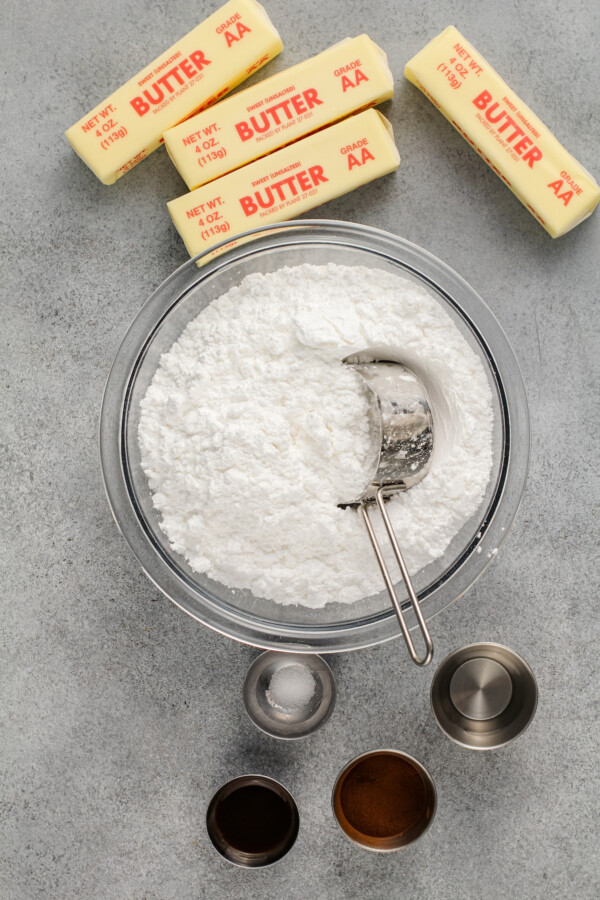 Sticks of butter and cups of vanilla extract and espresso are placed around a bowl of powdered sugar. 