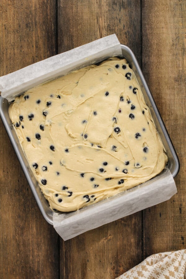 Overhead image of blueberry dough in a square baking pan with parchment paper hanging over the edges. 