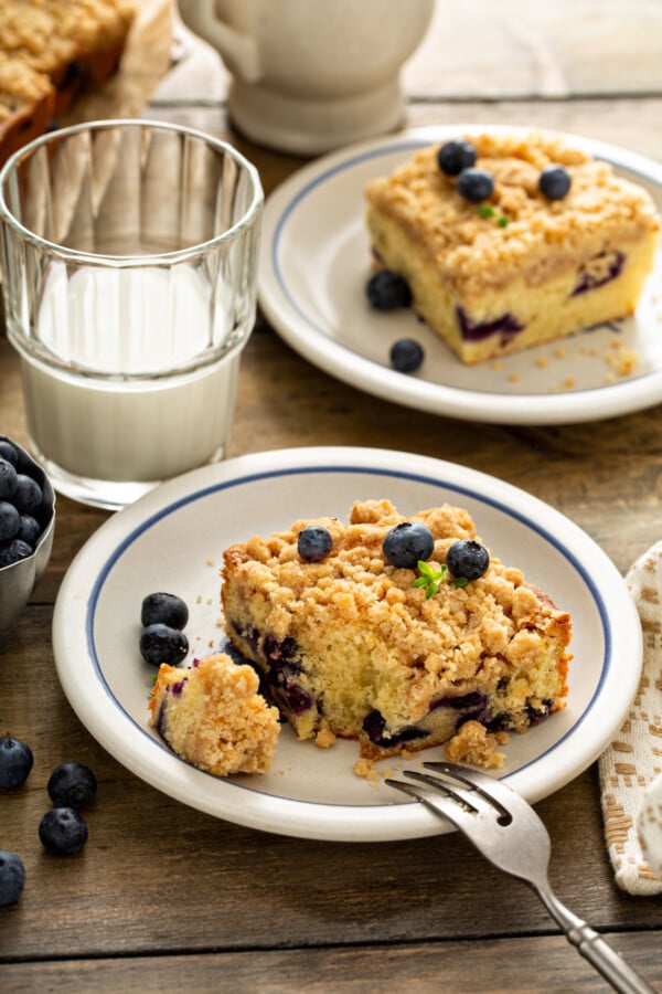 Coffee cake with blueberries on a white plate with a fork taking a bite out of it and a glass of milk. 