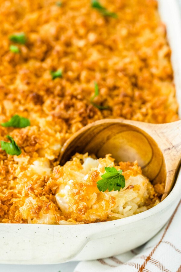 Up close image of cheese potato casserole in a white casserole dish with a wooden spoon taking a scoop. 