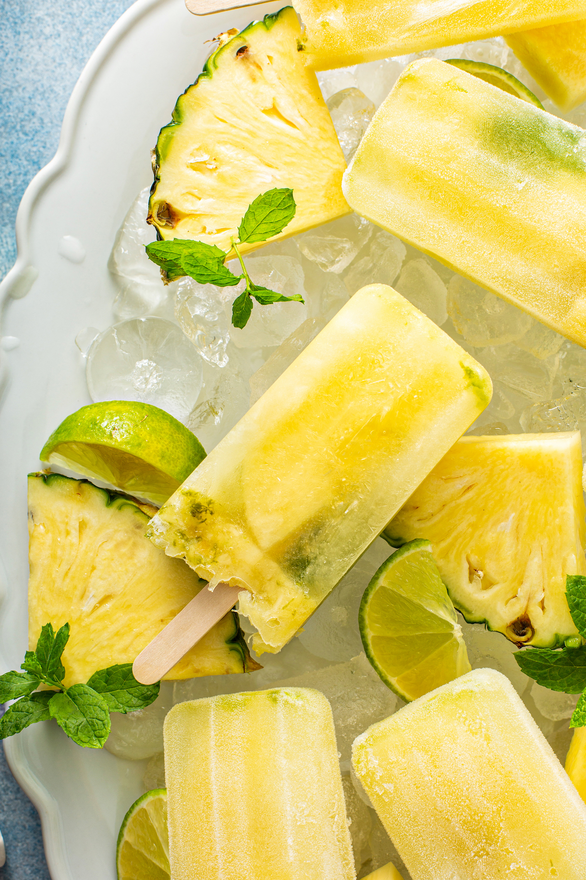 Up close image of pineapple popsicles on a platter with ice, pineapple wedges, lime and mint. 