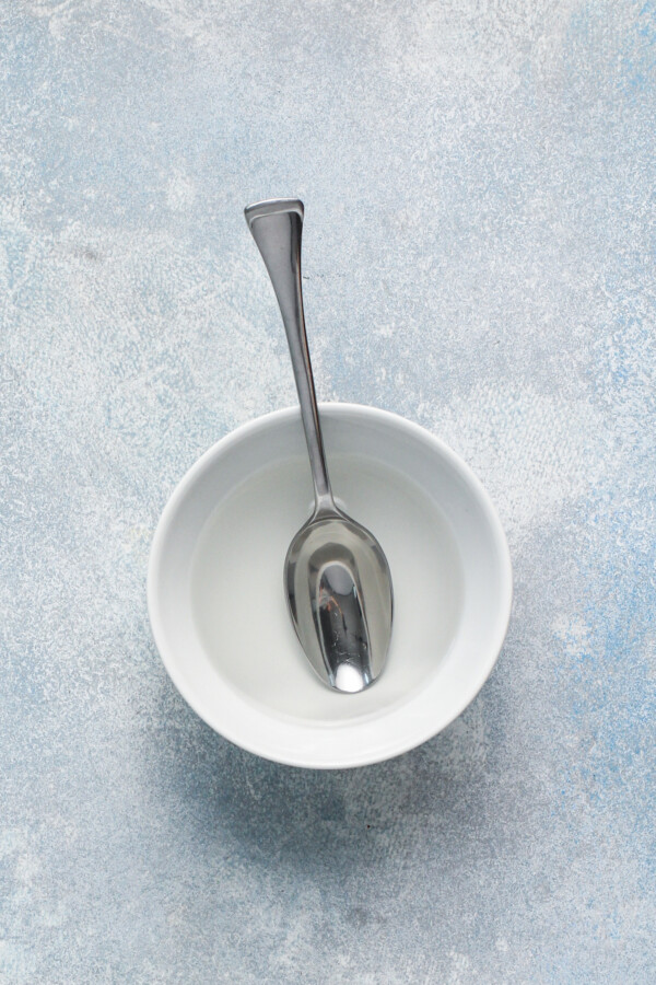 A white bowl with a metal spoon in it with water inside. 