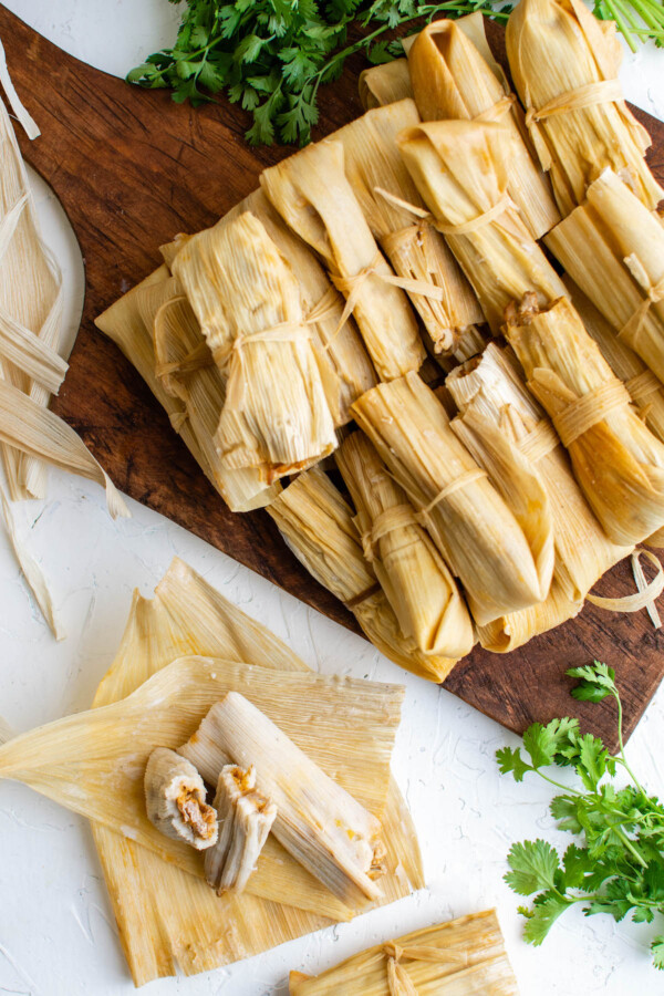 Overhead image of pork tamales all wrapped on a cutting board and one tamale opened to see the inside. 