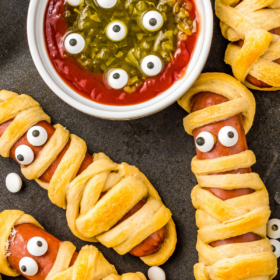 Overhead image of hot dogs wrapped with crescent dough baked with eye balls stuck on to look like a mummy.