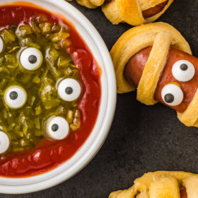 Mummy hot dogs on a baking sheet with a ketchup and relish dip with candy eyeballs.