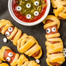 Mummy hot dogs on a black plate with a ketchup and relish dip with candy eyeballs.