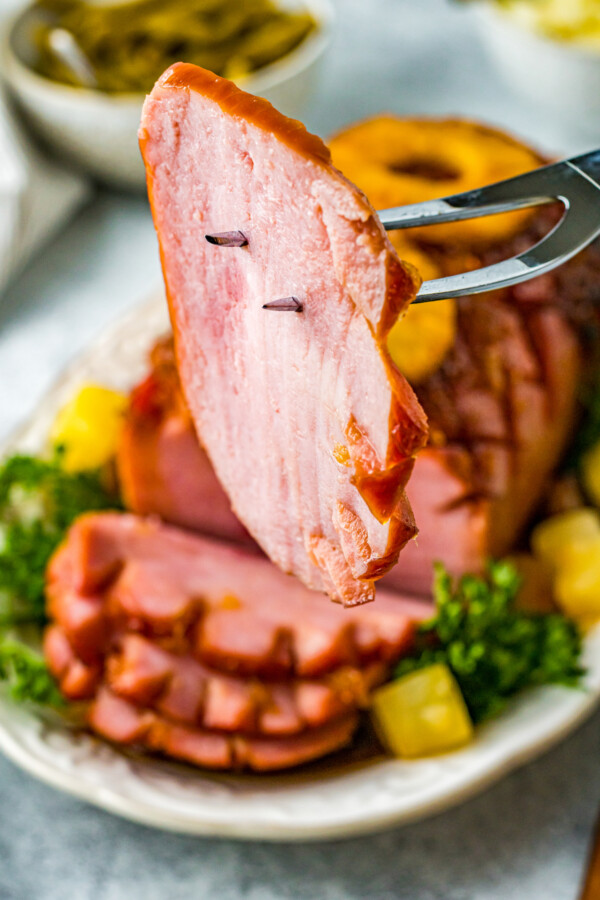 A close-up shot of a slice of air-fried ham on the tines of a serving fork, with the whole ham on a plate in the background, garnished with greenery and pineapple.