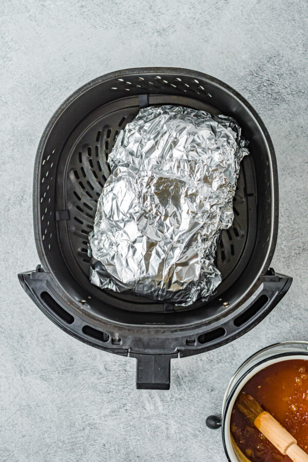 An air fryer basket with a foil wrapped ham inside.