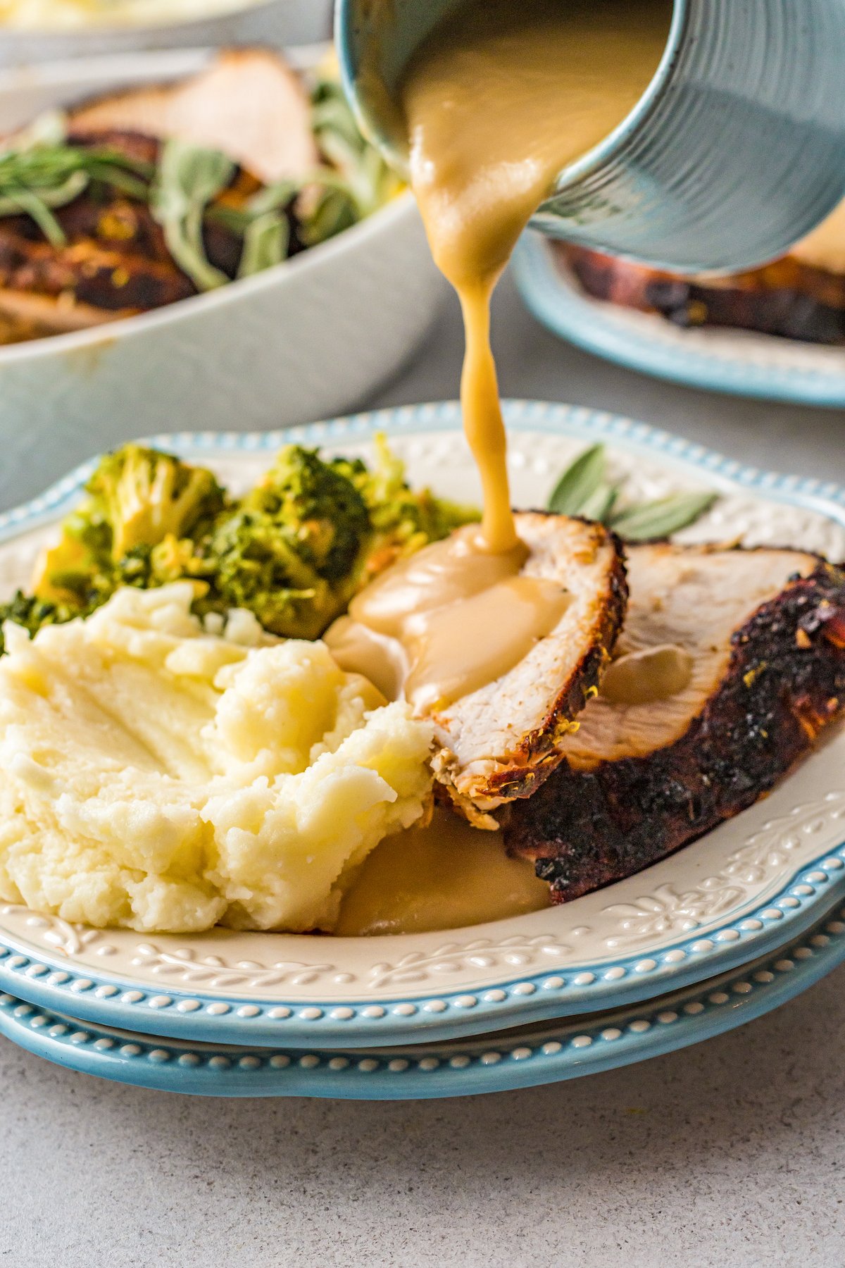 Gravy being poured over sliced turkey on a dinner plate with mashed potatoes and broccoli.