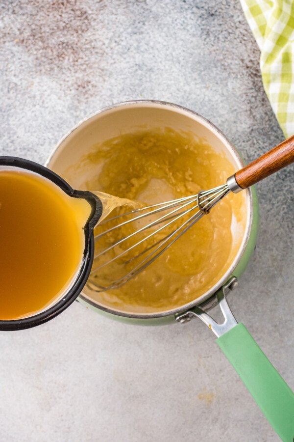 Chicken broth being poured over a roux in a small saucepan with whisk.