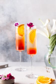 Aperol spritzes on a white background with a bouquet in the foreground of the shot. Each glass is decorated with small, edible purple flowers.